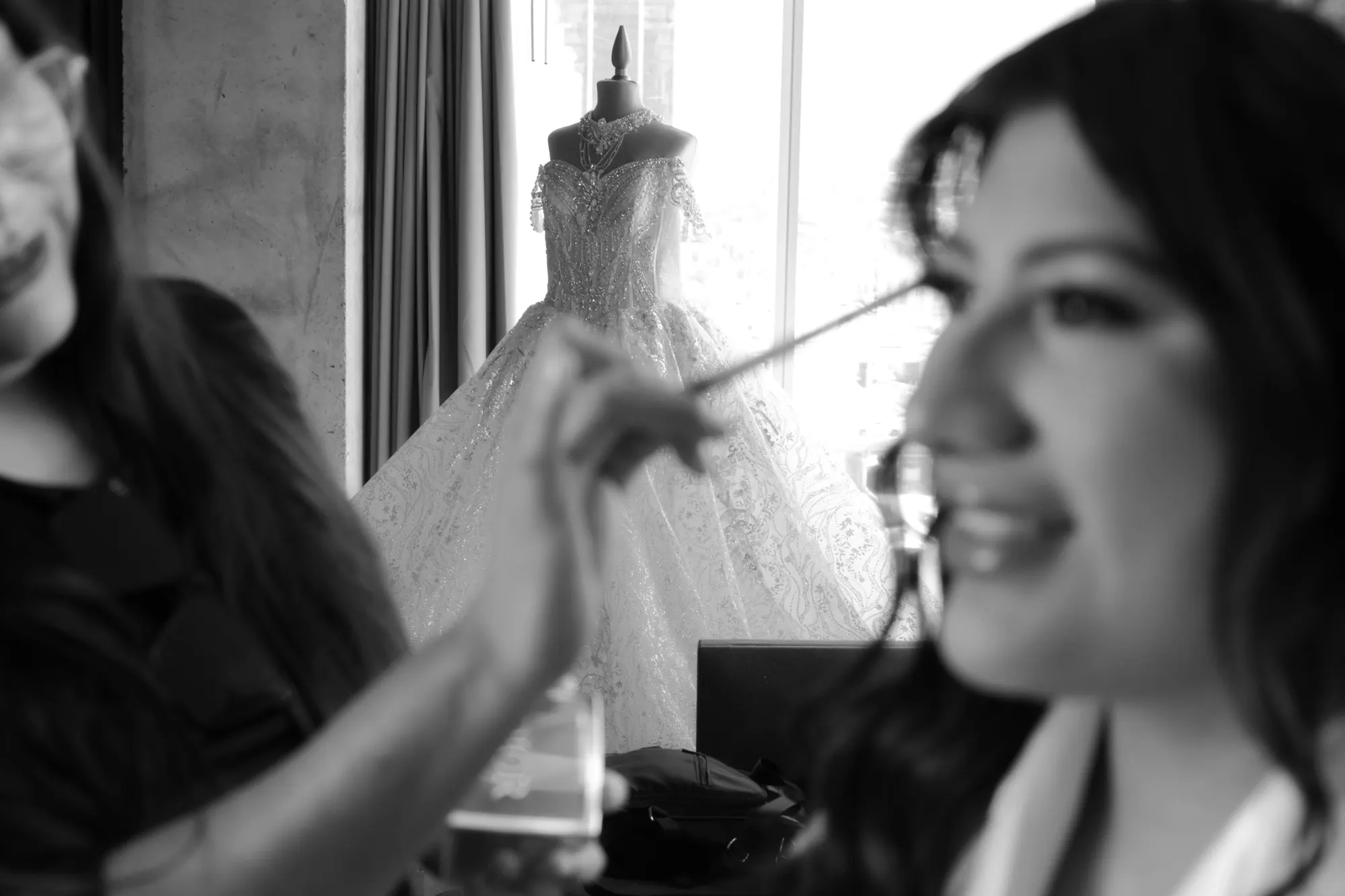 Novia maquillándose con el vestido al fondo, en blanco y negro