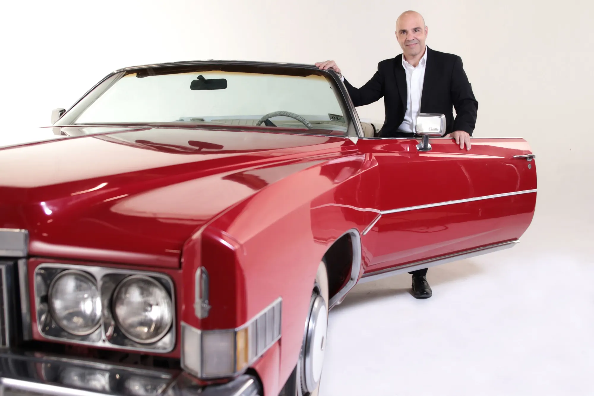 Hombre de traje junto a un convertible rojo clásico sobre fondo blanco