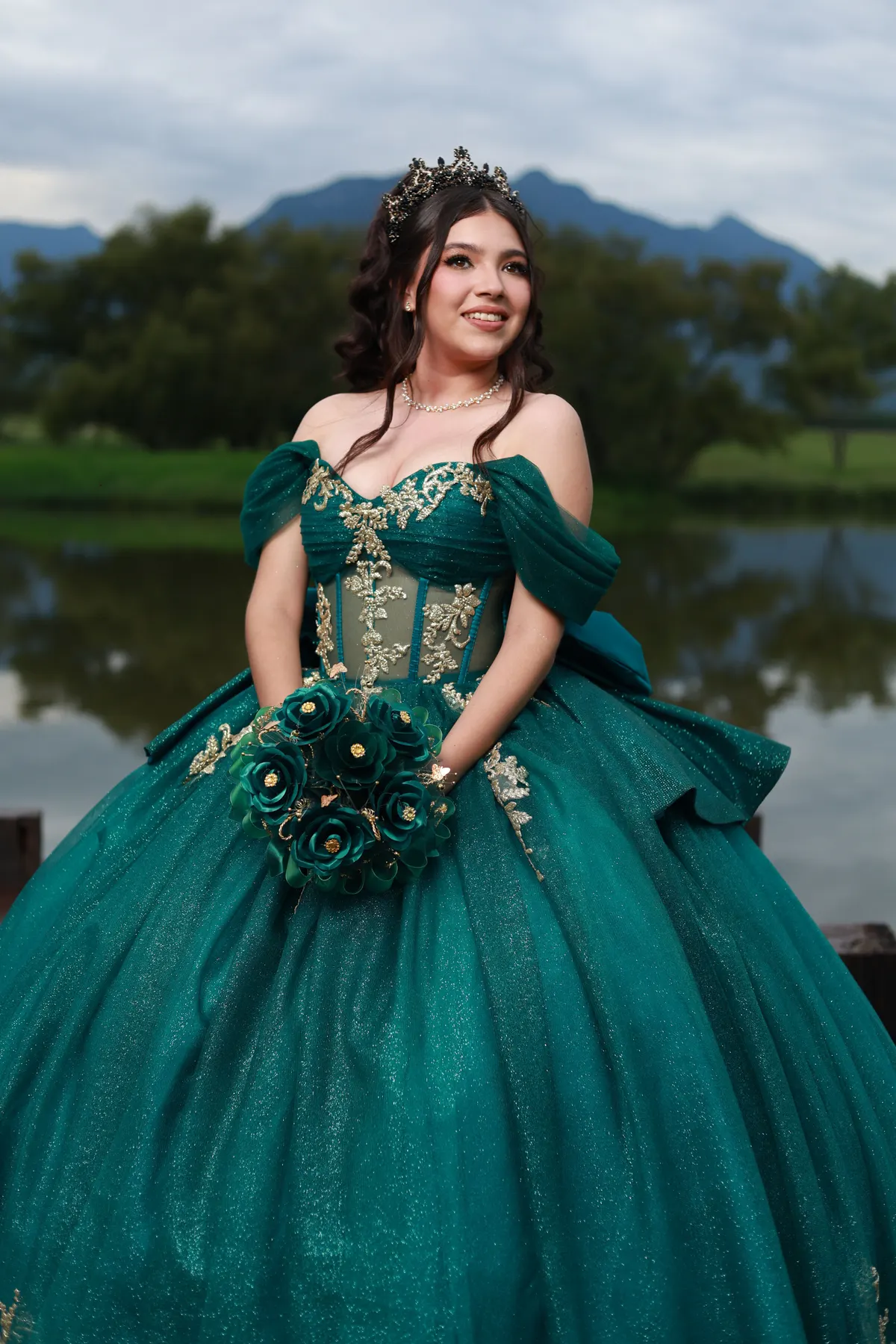 Quinceañera con vestido esmeralda junto a un lago con montañas al fondo